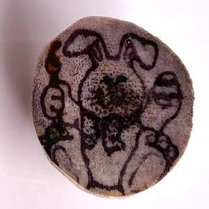 May include: A round, light-colored wooden slice with a hand-drawn illustration of a bunny holding an egg and a carrot. The bunny is depicted with dark outlines and a textured appearance, with a few green accents.