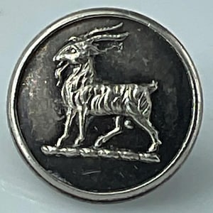 May include: A round, silver-toned button with a detailed relief of a goat. The goat is depicted in profile, with prominent horns and a beard, set against a dark background. The button has a raised edge.