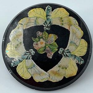 May include: A circular black brooch featuring a central floral cluster surrounded by three pale yellow and blue moths arranged in a ring. The moths have detailed wing patterns and are positioned with their bodies pointing toward the center flower.