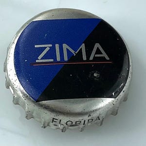 May include: A circular metal bottle cap with a silver-colored rim and a top surface divided diagonally into blue and black sections. The word ZIMA is printed in silver across the center, and the word FLORIIPA is embossed on the side of the cap.