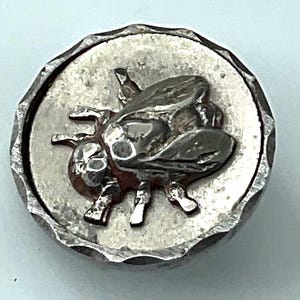 May include: A round, silver-toned button with a detailed relief of a bee. The bee is centered on a textured, circular background, surrounded by a textured, beveled edge. The button is likely made of metal.