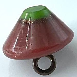 May include: A cone-shaped button with a green top and a reddish-brown body. The button has a metallic ring attached to the bottom. The button is likely made of glass or resin.