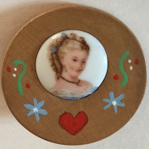 May include: A round wooden decorative piece featuring a painted portrait of a woman within a white circle. The wooden frame is adorned with painted blue flowers, a red heart, and green swirls with red dots. The portrait depicts a woman with blonde hair and a blue ribbon.