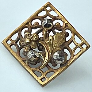 May include: A square-shaped gold-tone metal brooch featuring an openwork lattice frame. The center displays a floral motif with a textured gold flower, a textured gold leaf, and three small, faceted silver-tone metallic spheres.