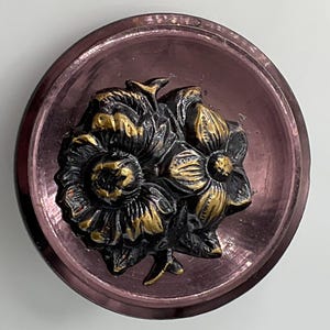May include: A vintage button with a raised floral design. The button has a dark purple, slightly iridescent background. The flowers are detailed with gold and dark purple accents, creating a striking contrast.