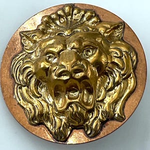 May include: A circular button featuring a high-relief, three-dimensional gold-toned lion head at the center. The lion has an open mouth, detailed mane, and expressive eyes. The background of the button is a flat, circular copper-toned metal disc.