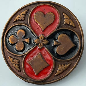May include: A round metal button featuring a raised design of the four playing card suits. The heart and diamond are set against a red background, while the club and spade are set against a black background. A small floral motif sits in the center.