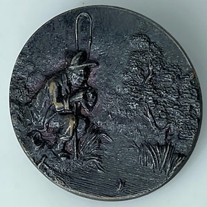 May include: A round, dark metal button with a detailed relief of a fisherman seated near a tree. The fisherman wears a hat and holds a fishing rod. The scene is set in a natural environment with tall grass and foliage.