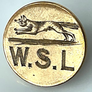 May include: A gold-colored, circular pin with a detailed engraving of a running fox above the letters "W.S.L." The fox is depicted in motion, with its legs extended. The pin has a slightly worn appearance.
