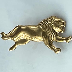 May include: A gold-toned metal brooch shaped like a lion in a running or leaping pose. The lion is depicted in profile with its mouth open, showing teeth, and features detailed engravings on its mane and muscular body, capturing a sense of motion.
