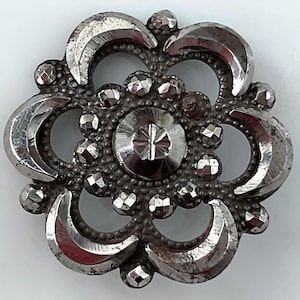 May include: A circular silver-toned metal button featuring a central faceted circular element surrounded by six crescent-shaped segments and small, textured, faceted bead-like accents arranged in a floral pattern with a dark, aged patina.