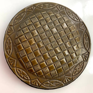 May include: A round, dark brown, ceramic plate with a raised, checkered pattern. The plate has a decorative border with a repeating leaf design.