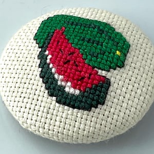 May include: A round, off-white fabric button with a cross-stitch design of a watermelon slice. The watermelon features red flesh, a black rind, and green and black details. The button is approximately 2 inches in diameter.