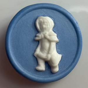 May include: A blue ceramic button with a white raised image of a girl wearing a hooded cloak and carrying a bucket.