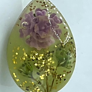 May include: A teardrop-shaped resin pendant featuring a translucent light green base embedded with a purple flower, green foliage, and scattered gold glitter flakes. A small metal jump ring is attached near the top of the pendant.