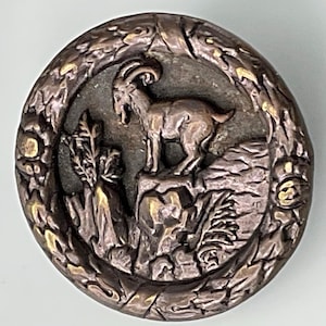 May include: A round, bronze-colored button with a raised image of a mountain goat standing on a rock. The goat has a long, curved beard and horns. The button is surrounded by a decorative border.