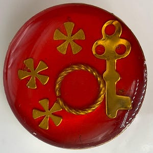 May include: A round, red decorative button with gold accents. The button features a gold key, three gold starbursts, and a gold rope circle. The button has a glossy finish.