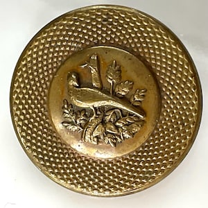 May include: A round, antique brass button with a textured outer ring and a central design. The center features a detailed relief of a bird, possibly a pheasant, amidst foliage. The button is likely for sewing or decorative purposes.