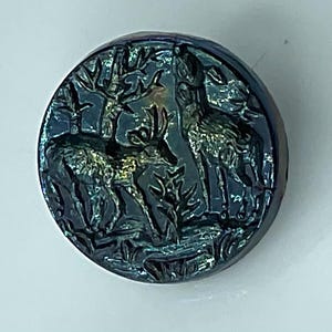 May include: A round, iridescent button with a detailed relief of two deer in a forest setting. The button has a dark blue and green color scheme, with highlights that catch the light. The design includes trees and foliage.