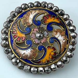 May include: A round, antique brooch with a detailed floral design. The center features a pink flower with a silver-colored gem. The brooch has a blue, yellow, red, and brown enamel design, surrounded by a border of silver-colored beads.