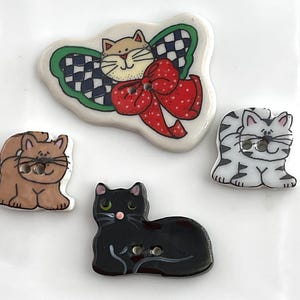 May include: A collection of decorative cat-themed buttons. The buttons feature various cat designs, including a brown cat, a black cat, a white cat with gray stripes, and a cat with a bow. The buttons are in various shapes and sizes.