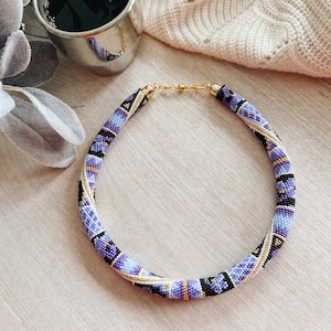 Könnte beinhalten: Eine violette, schwarze und goldene Perlenkette mit geometrischem Muster. Die Kette besteht aus kleinen Perlen und hat einen Verschluss.