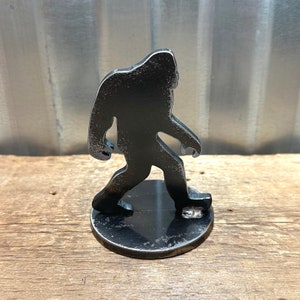 May include: A black metal silhouette of Bigfoot walking, standing on a round black metal base.
