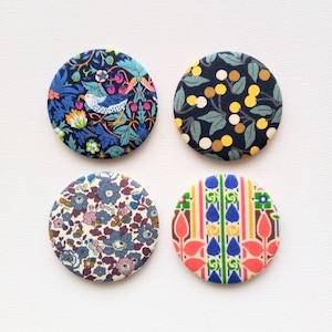 May include: Four round fabric-covered buttons featuring different floral and geometric patterns. The buttons are in shades of blue, green, yellow, red, and white.