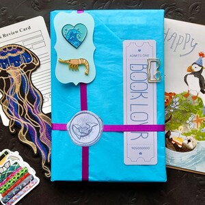 Cita a ciegas con un libro – Caja de regalo edición playa de verano con pins, pegatinas, tarjeta y sorpresas / Regalo misterioso para amantes de los libros con temática oceánica