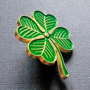 Puede incluir: Un broche de trébol de cuatro hojas con borde dorado y relleno de esmalte verde brillante. El broche tiene un diseño detallado con vetas visibles en cada hoja. El trébol es un símbolo de buena suerte, sobre un fondo texturizado.