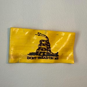 May include: A yellow flag with a black coiled snake and the words "DON'T TREAD ON ME". The flag is rectangular and has a slightly textured surface. The snake is in the center of the flag.
