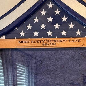 May include: A wooden flag display case with a blue and white star-spangled background. The case has a wooden plaque with the inscription "MSGT RUSTY "LUXURY" LANE 1988-2008". The case is designed to hold a folded American flag.