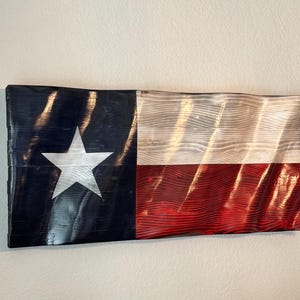 May include: Wooden Texas flag wall art. The flag features a large white star on a navy blue background, with a white and red striped section. The wood grain is visible, adding texture to the piece. A decorative item for home decor.