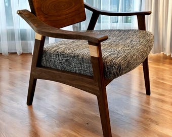 May include: A mid-century modern armchair with a dark wood frame and a patterned gray and white fabric seat. The chair has a curved back and armrests, with light-colored wood accents at the joints. The chair is set on a hardwood floor.