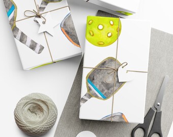 May include: Three white gift boxes with a watercolor design of pickleball paddles and balls. The boxes are tied with twine and have white gift tags attached.