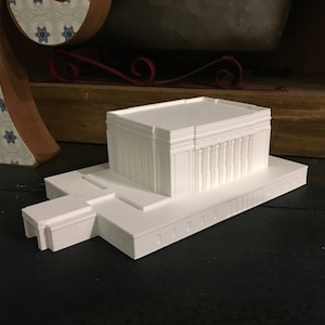 May include: White 3D printed model of a building with a flat roof and a small entrance. The model is on a black surface.