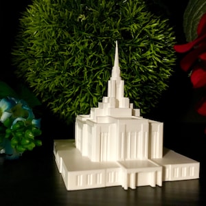 May include: A white miniature replica of a Mormon temple with a tall spire and intricate details. The temple is sitting on a dark surface with green and red plants in the background.