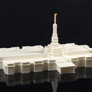 May include: A white 3D printed model of a temple with a gold statue on top of the spire.