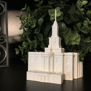 May include: White miniature replica of a temple with a tall spire and intricate details. The model is on a black surface with green foliage in the background.