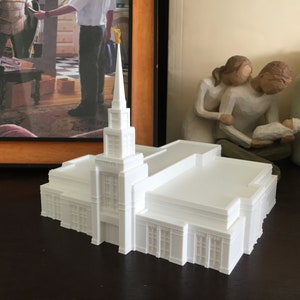 May include: A white 3D printed model of a church with a tall spire and intricate details. The model is sitting on a dark wooden surface.