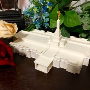 May include: A white 3D printed model of a temple with a gold spire. The model is sitting on a wooden surface with a red rose and a cream-colored rose in the foreground.