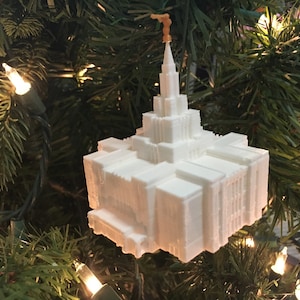 May include: A white temple-shaped Christmas tree ornament, with a detailed, layered design and a spire topped with a small, gold-coloured figure. The ornament hangs on a Christmas tree decorated with fairy lights.