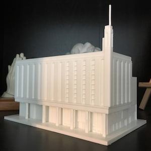 May include: White 3D printed model of a building with a tall spire. The building has a rectangular shape with multiple windows and a flat roof.