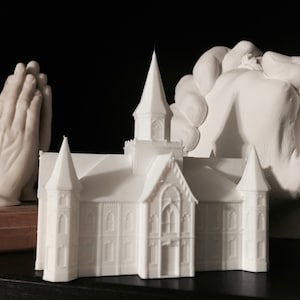 May include: A white 3D printed model of a church building with two towers and a spire. The model is on a black surface next to two white 3D printed hands in a praying position.