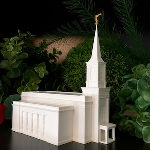 May include: A white model church with a tall spire and a gold-coloured weather vane. The building has a rectangular base with vertical lines and is surrounded by green foliage.