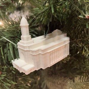 May include: White ceramic ornament shaped like a church building with a steeple. The ornament is hanging from a Christmas tree.