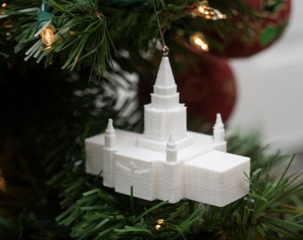 Op de afbeelding: Witte 3D-geprinte ornament van een kerkgebouw, hangend aan een kerstboom met groene naalden en heldere kerstverlichting.