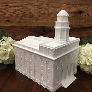 Op de afbeelding: Wit 3D-geprint model van een tempel met een toren op de top. Het model staat op een houten oppervlak met witte bloemen aan beide kanten.