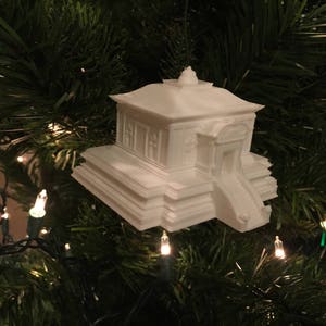 Op de afbeelding: Witte keramische ornament in de vorm van een gebouw met een trapsgewijs dak en treden. De ornament hangt aan een kerstboom.