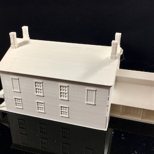 Op de afbeelding: Een wit 3D-geprint model van een huis met twee verdiepingen, een veranda en meerdere ramen.